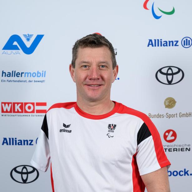 Oliver Dreier: Ein Mann im rot-weißen Sporttrikot mit österreichischem Wappen steht vor einer Sponsorenwand und lächelt.