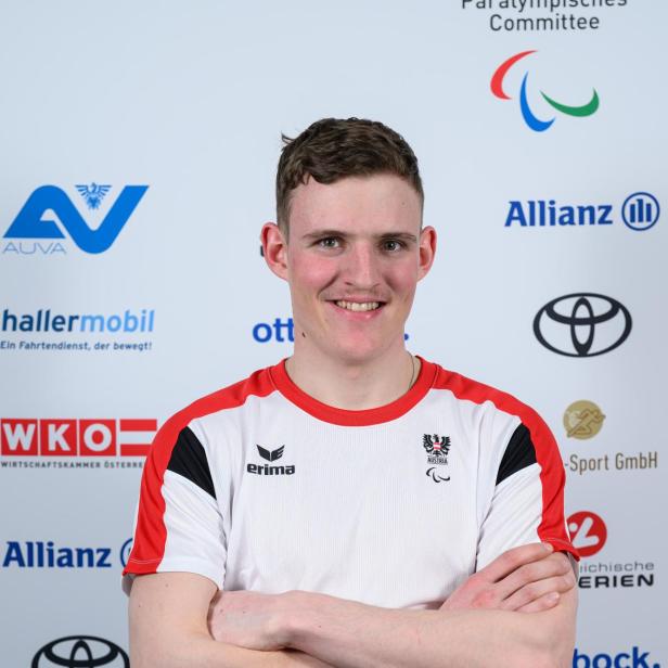 Johannes Aigner: Junger Mann im weißen Sporttrikot mit roten Akzenten steht lächelnd mit verschränkten Armen vor Sponsorenlogos.