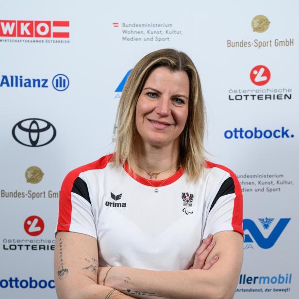 Christina Obwexer: Eine Frau im Rollstuhl mit verschränkten Armen trägt ein Sporttrikot und lächelt vor einer Sponsorenwand.