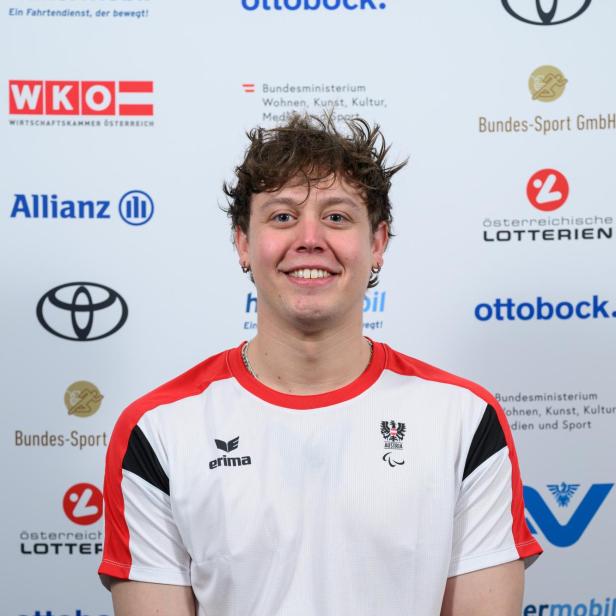 Sebastian Rigby: Junger Mann im Rollstuhl mit kurzem, lockigem Haar trägt ein weißes Sporttrikot mit roten Akzenten und lächelt vor Sponsorenlogos.