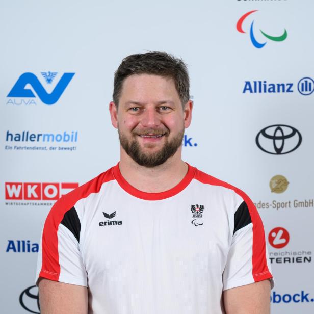 Michael Scharnagl: Ein Mann mit Bart trägt ein weißes Sportshirt mit roten Akzenten und steht vor einer Sponsorenwand.