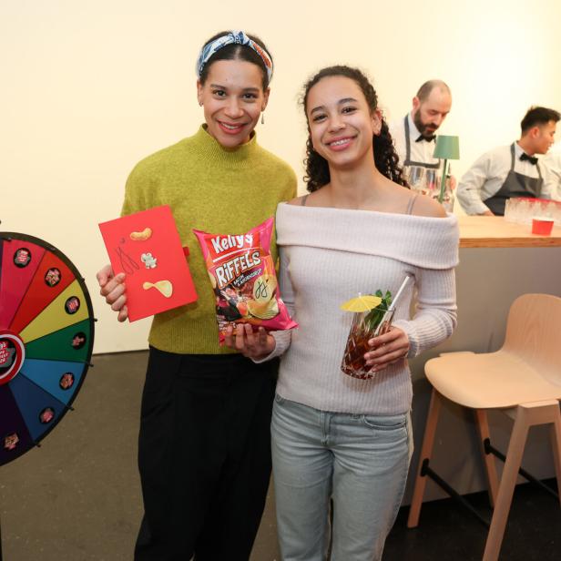 Zwei junge Frauen stehen lächelnd vor einer Bar, eine hält Chips und eine Karte, die andere ein Getränk mit Schirmchen.