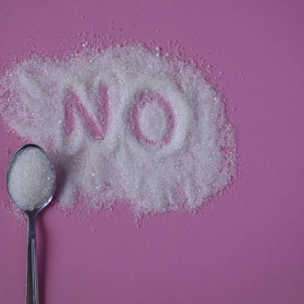 Ein Löffel Zucker liegt neben einem Haufen Zucker, in den das Wort „NO“ geschrieben ist, auf rosa Hintergrund.