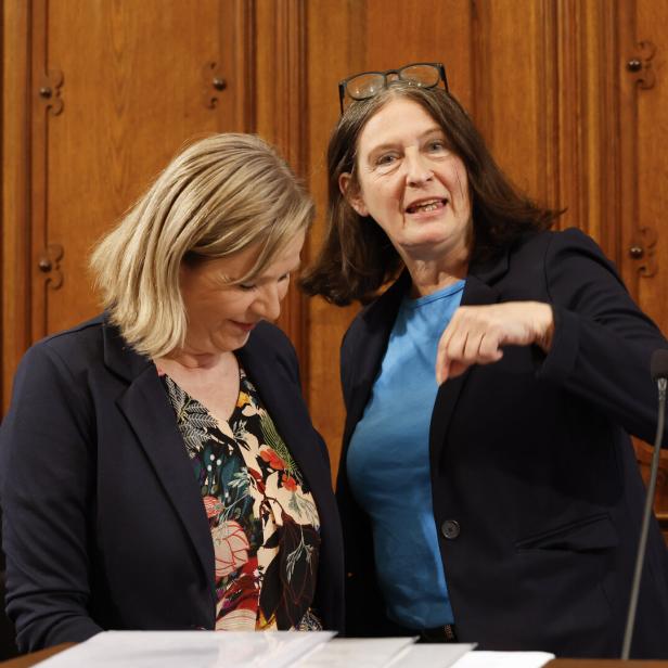 Zwei Frauen in Anzügen stehen vor einer hölzernen Wand und diskutieren.