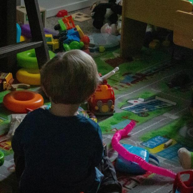 Ein kleiner Junge sitzt in einem unordentlichen, mit Spielzeug übersäten Kinderzimmer auf dem Boden.