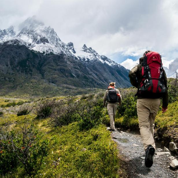 Zwei Wanderer mit Rucksäcken gehen auf einem schmalen Bergpfad vor schneebedeckten Gipfeln durch eine alpine Landschaft.