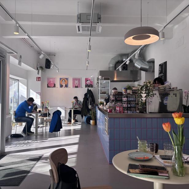 Helles Café mit wenigen Gästen, moderner Einrichtung, Blumen auf den Tischen und bunten Porträts an der Wand.