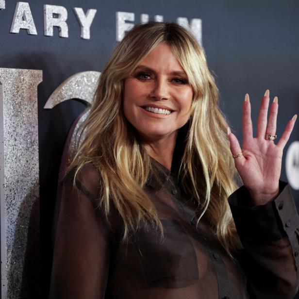 Heidi Klum lächelt und winkt vor einem dunklen Hintergrund mit silbernen Schriftzügen.