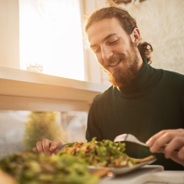 Ein Mann mit Bart sitzt am Tisch und isst bei Sonnenlicht einen Teller mit frischem Salat.