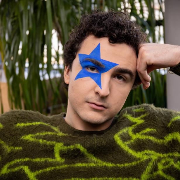 Eine Person mit lockigen Haaren trägt einen grünen Pullover und hat einen großen blauen Stern um das rechte Auge gemalt.