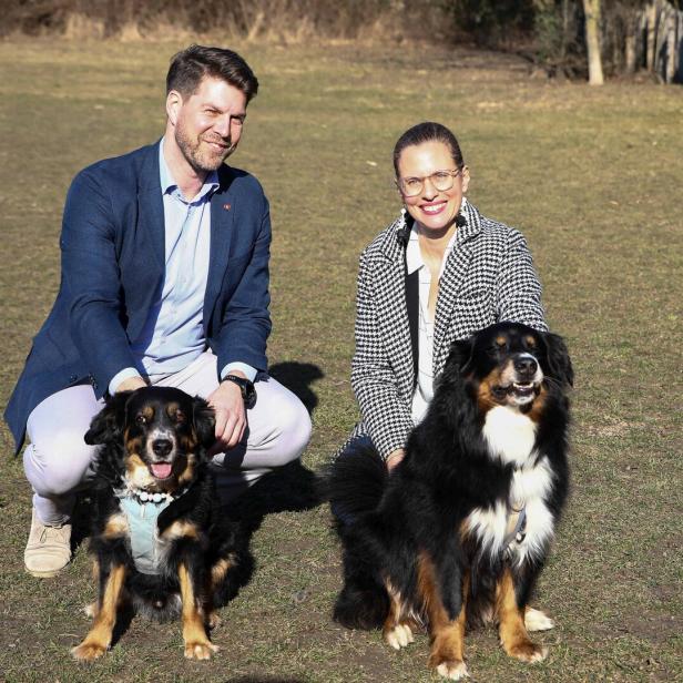 Ein Mann und eine Frau knien auf einer Wiese und lächeln, neben ihnen sitzen zwei große, schwarz-braune Hunde.