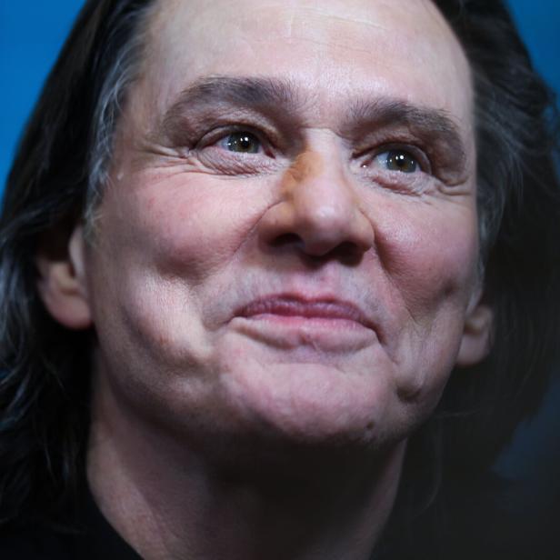 Jim Carrey lächelt mit leicht geneigtem Kopf vor einem blauen Hintergrund.