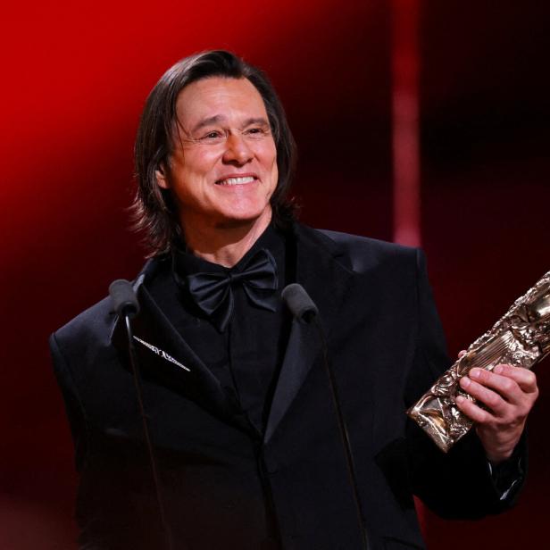 Jim Carreyim schwarzen Anzug mit Fliege hält eine goldene Trophäe und lächelt vor rotem Hintergrund.