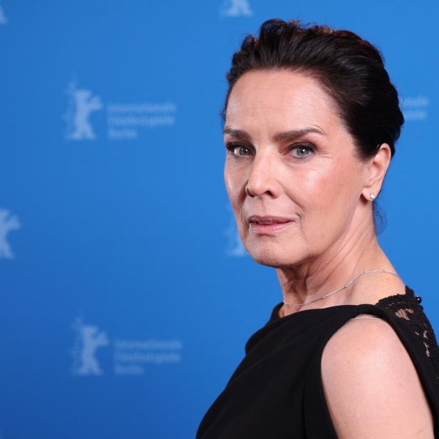 Désirée Nosbusch  mit dunklem Haar und schwarzem Kleid posiert vor einem blauen Hintergrund.