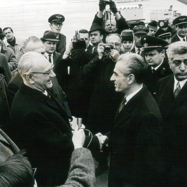 Mehrere Männer in Anzügen und Uniformen begrüßen sich auf einem Flughafen, umgeben von Fotografen und Journalisten.