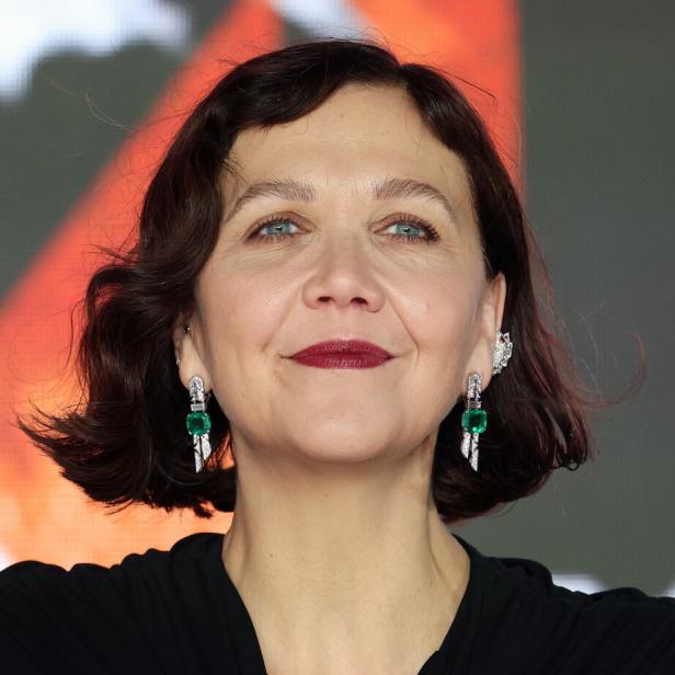 Maggie Gyllenhaal lächelt mit dunklem Lippenstift und smaragdgrünen Ohrringen.