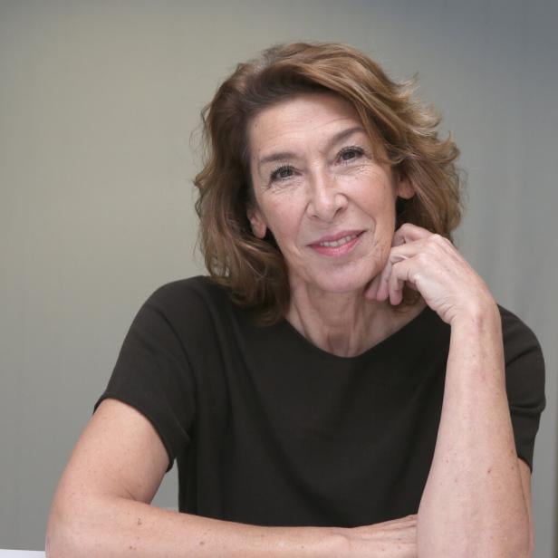 Adele Neuhauser mit braunem Haar und dunklem Oberteil lächelt, den Kopf auf ihre Hand gestützt.