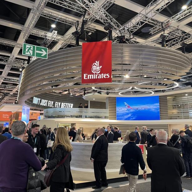 Viele Menschen stehen an einem modernen Messestand von Emirates mit großem Logo und Bildschirm, der ein Flugzeug zeigt.
