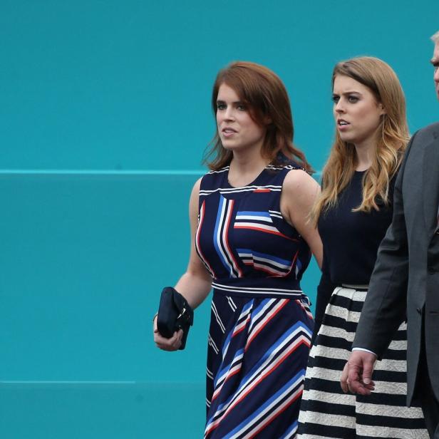 Prinz Andrew geht mit Prinzessin Beatrice und Prinzessin Eugenie vor einem türkisfarbenen Hintergrund.