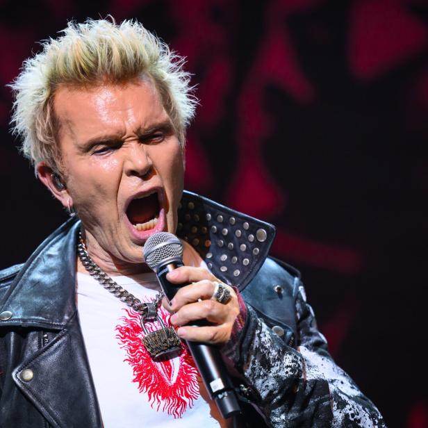 Billy Idol singt mit offenem Mund in ein Mikrofon, bekleidet mit Lederjacke und Kette.