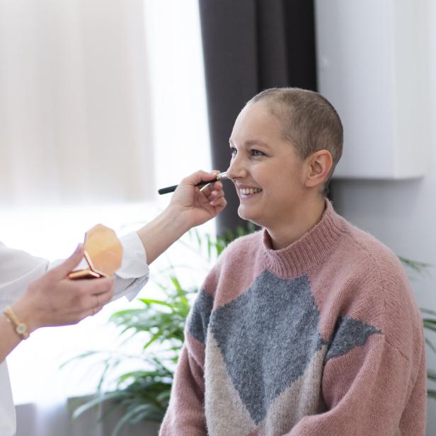 Eine Frau trägt einer lächelnden Person mit sehr kurzen Haaren Make-up mit einem Pinsel auf.