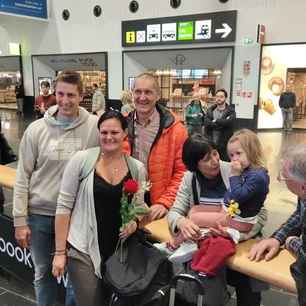Sechs Menschen, darunter ein kleines Kind, stehen und sitzen lächelnd zusammen in einer belebten Bahnhofshalle.