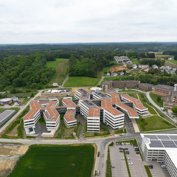 Mehrere moderne Gebäude mit roten Dächern und großen Fenstern liegen inmitten grüner Landschaft und Waldflächen.