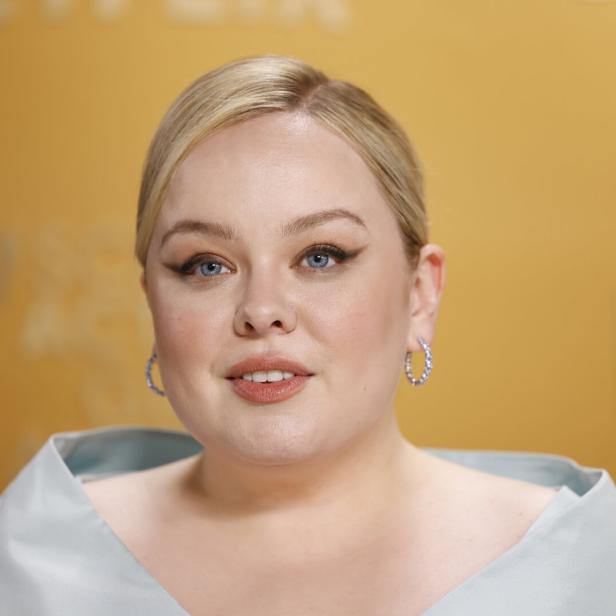 Nicola Coughlan mit blonden Haaren und hellblauen Augen lächelt vor einem gelben Hintergrund.