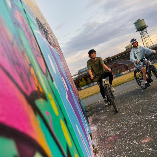zwei Personen fahren mit dem Fahrrad neben einer Wand, die mit Graffiti bemalt ist