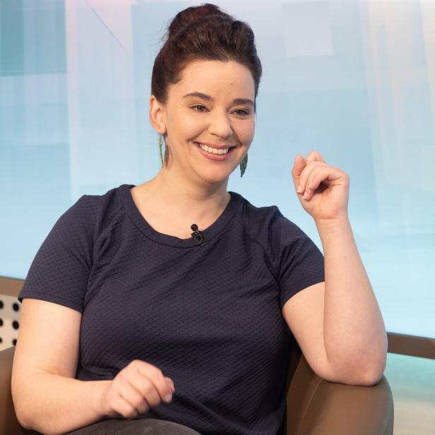 Eine Frau mit dunklen Haaren sitzt lächelnd auf einem Sessel und trägt ein dunkelblaues Oberteil.