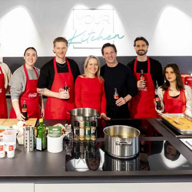 Acht Personen mit roten Coca-Cola-Schürzen stehen lächelnd in einer modernen Küche.