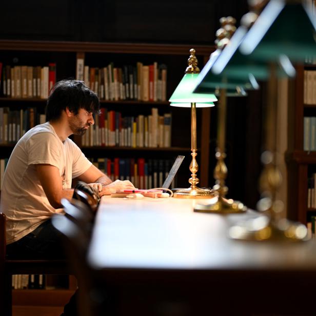 Ein Mann sitzt an einem langen Tisch in einer Bibliothek und arbeitet an einem Laptop unter einer grünen Lampe.