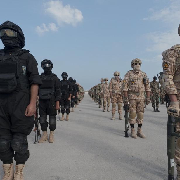 Mehrere Soldaten in unterschiedlichen Uniformen und Ausrüstungen stehen in Reihen auf einer Straße unter blauem Himmel.
