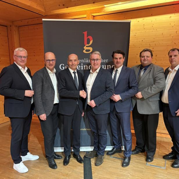 Sieben Männer in Anzügen stehen nebeneinander in einem holzvertäfelten Raum und posieren vor einem schwarzen Banner.