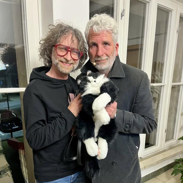 Zwei Männer stehen nebeneinander und halten gemeinsam eine schwarz-weiße Plüschkatze in den Armen.