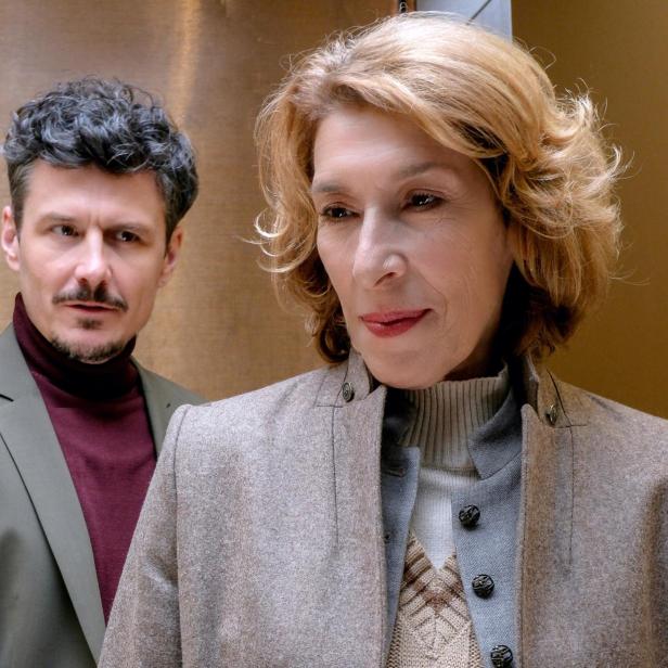 Eine Frau mit blondem Haar steht im Vordergrund, ein Mann mit dunklem Haar blickt sie ernst von hinten an.