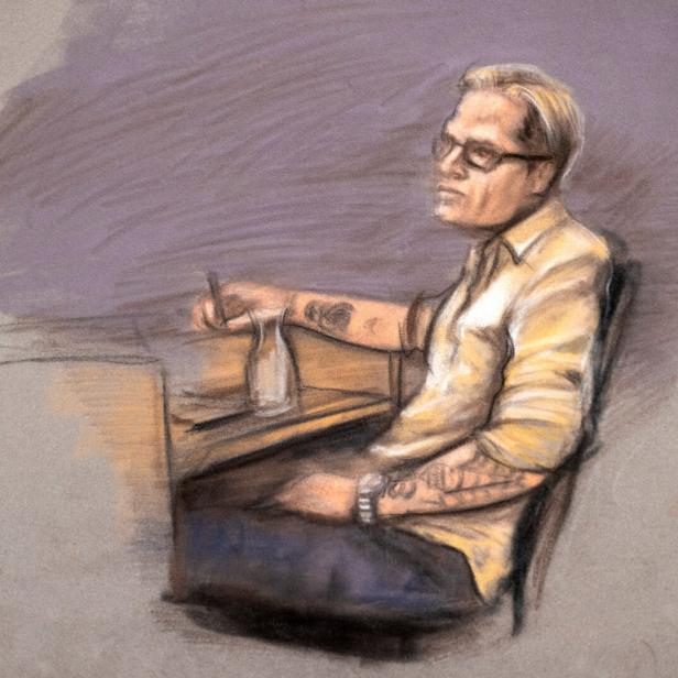 Eine Person mit Brille und sichtbaren Tattoos sitzt an einem Tisch und hält ein Glas mit Strohhalm.