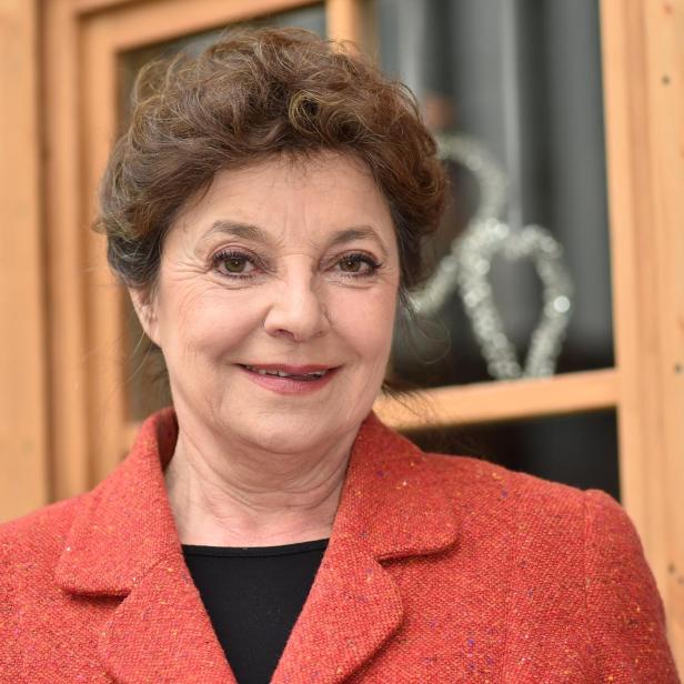 Eine lächelnde Frau mit braunem Haar trägt ein rotes Jackett vor einem Fenster.