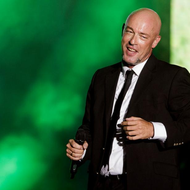 Der Graf von Unheilig steht im schwarzen Anzug mit Mikrofon auf einer Bühne vor grünem Hintergrund.