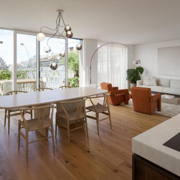 Helles, modern eingerichtetes Wohnzimmer mit großem Esstisch, beigen Sofa, zwei orangefarbenen Sesseln und großen Fenstern.