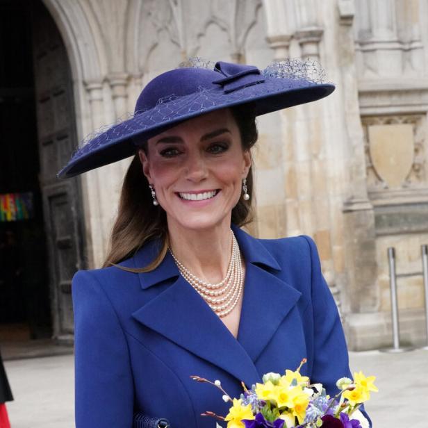 Kate trägt einen blauen Hut und Blazer und hält einen Blumenstrauß.