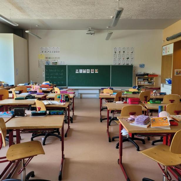 Mehrere Schultische und Stühle stehen in einem hellen Klassenraum, auf den Tischen liegen Schulmaterialien und Hefte.