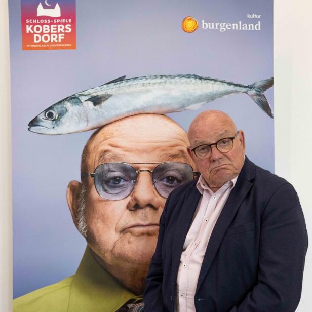 Ein älterer Mann mit Brille steht vor einem Plakat, das sein Porträt mit einem Fisch auf dem Kopf zeigt.
