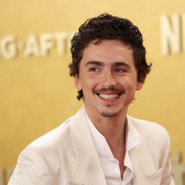 Ein lächelnder Timothée Chalamet mit dunklen Locken und weißem Anzug vor goldenem Hintergrund.
