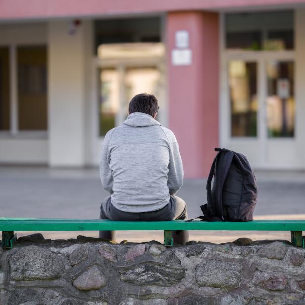 One young man sitting on bench at school yard. Break time. Back view.