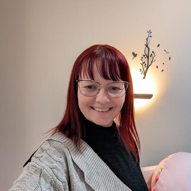 Frau mit Brille und roten Haaren lächelt vor heller Wand mit Wanddeko und Lampe.