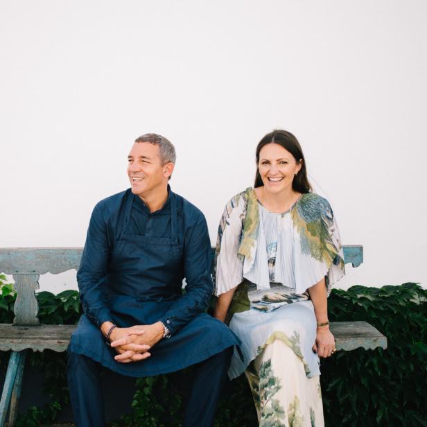 Ein Mann und eine Frau sitzen lachend auf einer alten Holzbank vor einer weißen Wand mit grünen Pflanzen im Hintergrund.