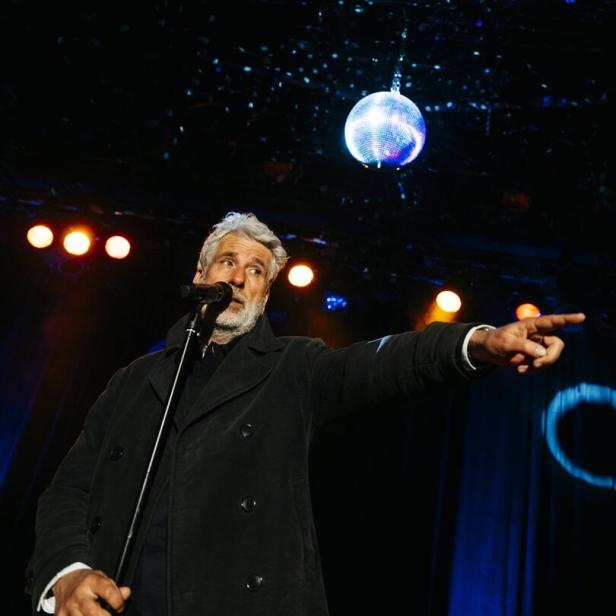 Ein Mann mit grauem Haar spricht in ein Mikrofon und zeigt auf eine Bühne, über ihm hängt eine leuchtende Discokugel.