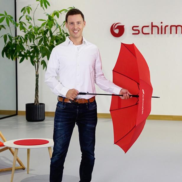 Ein Mann mit einem roten Schirm steht vor dem Schild "Schirmmacher"