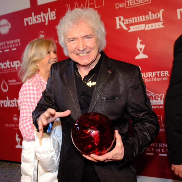 Ein Mann mit grauen Haaren hält eine rote Bowlingkugel vor einem roten Hintergrund mit Logos.